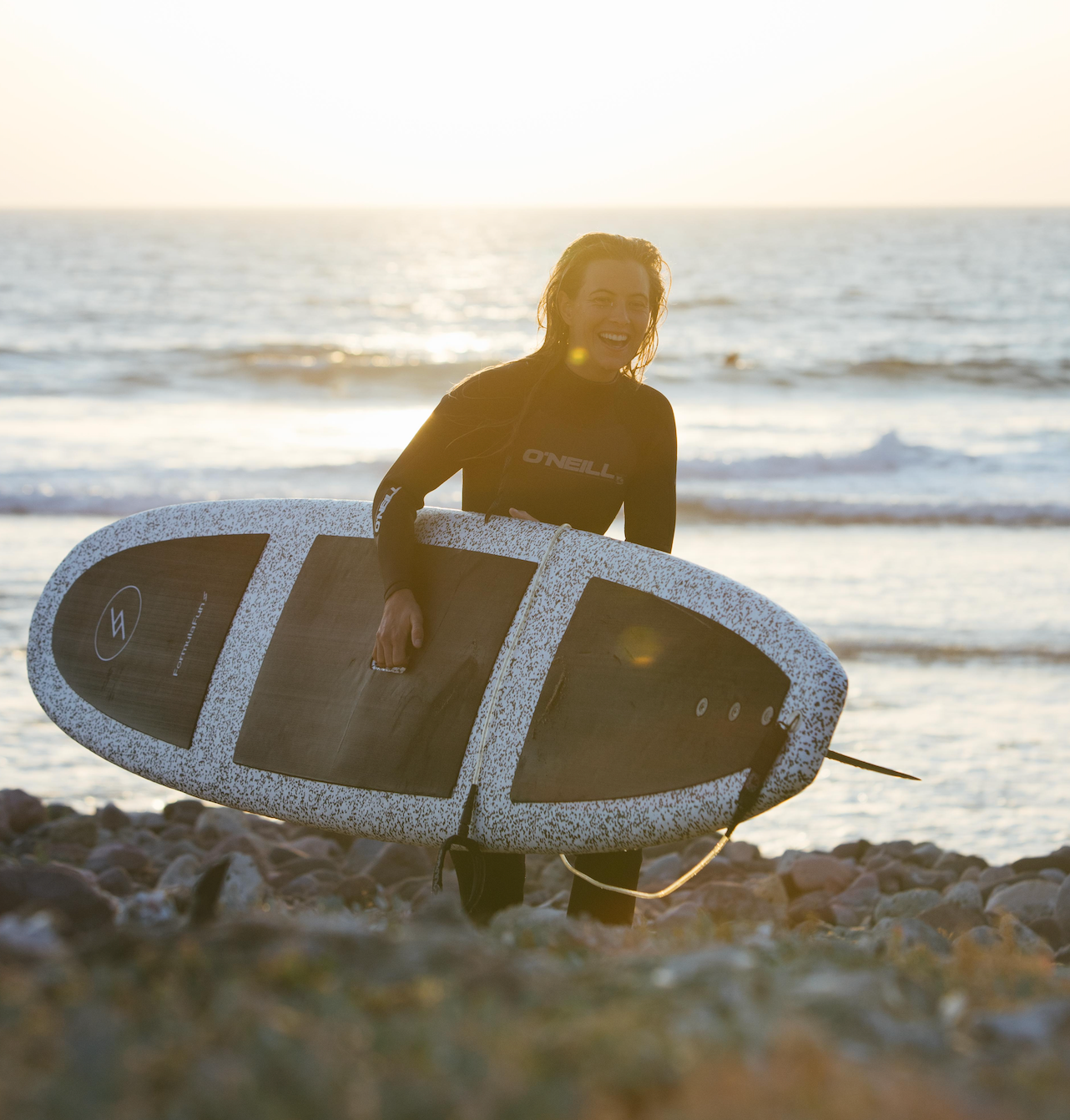 Woman on a beach at sunset carrying a speckled Formula Fun Foamies surfboard