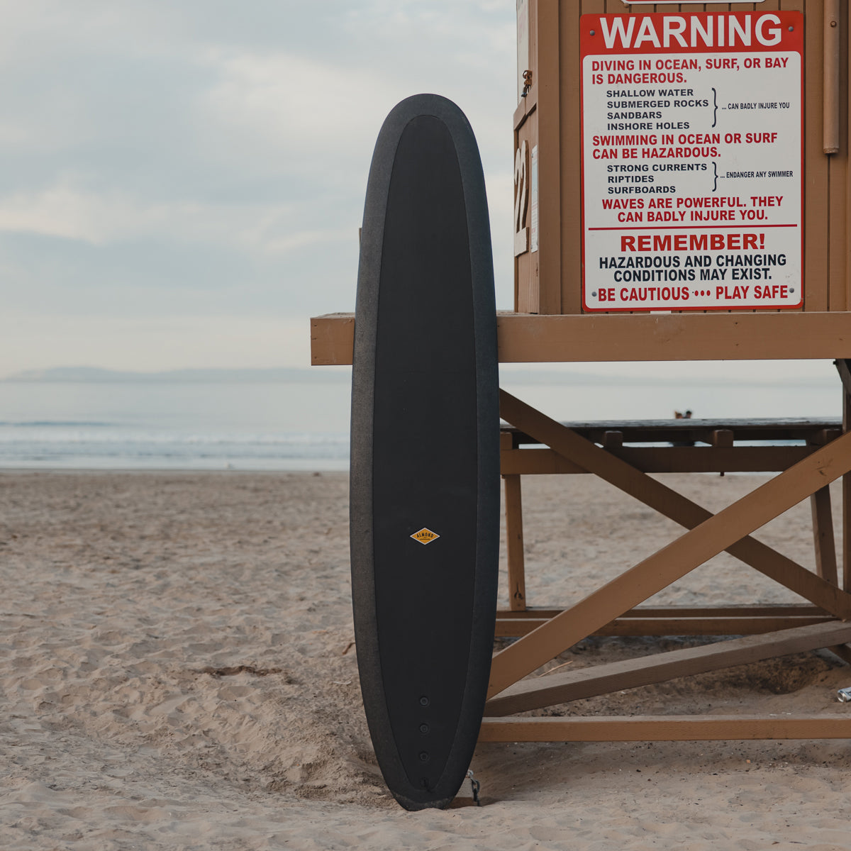 A black 9 foot 2 inch Almond Surf Thump surfboard leaning up against a lifeguard tower on a beach