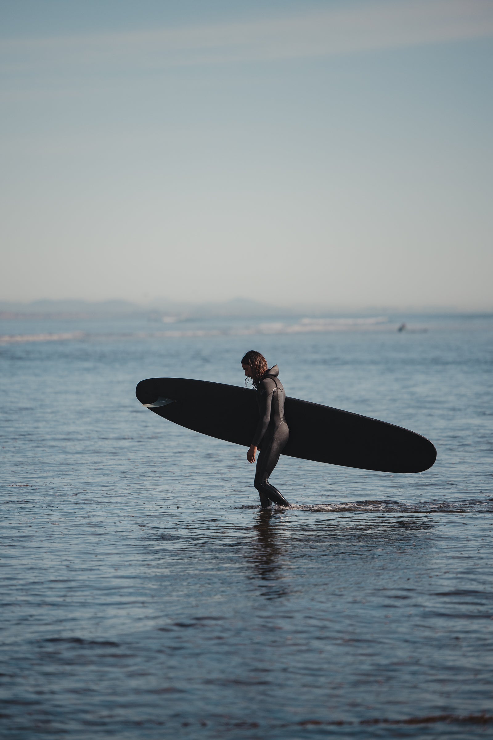 Man entering the ocean in a wetsuit with a black 9 foot 2 inch Almond Surf Thump surfboard