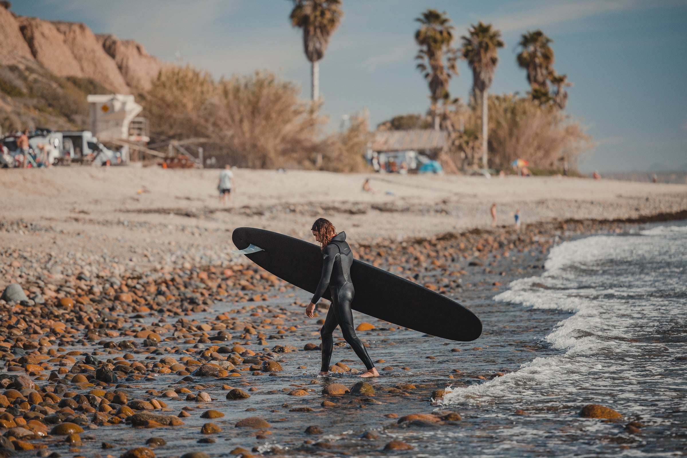 Man coming out of the ocean onto the beach in a wetsuit with a black 9 foot 2 inch Almond Surf Thump surfboard