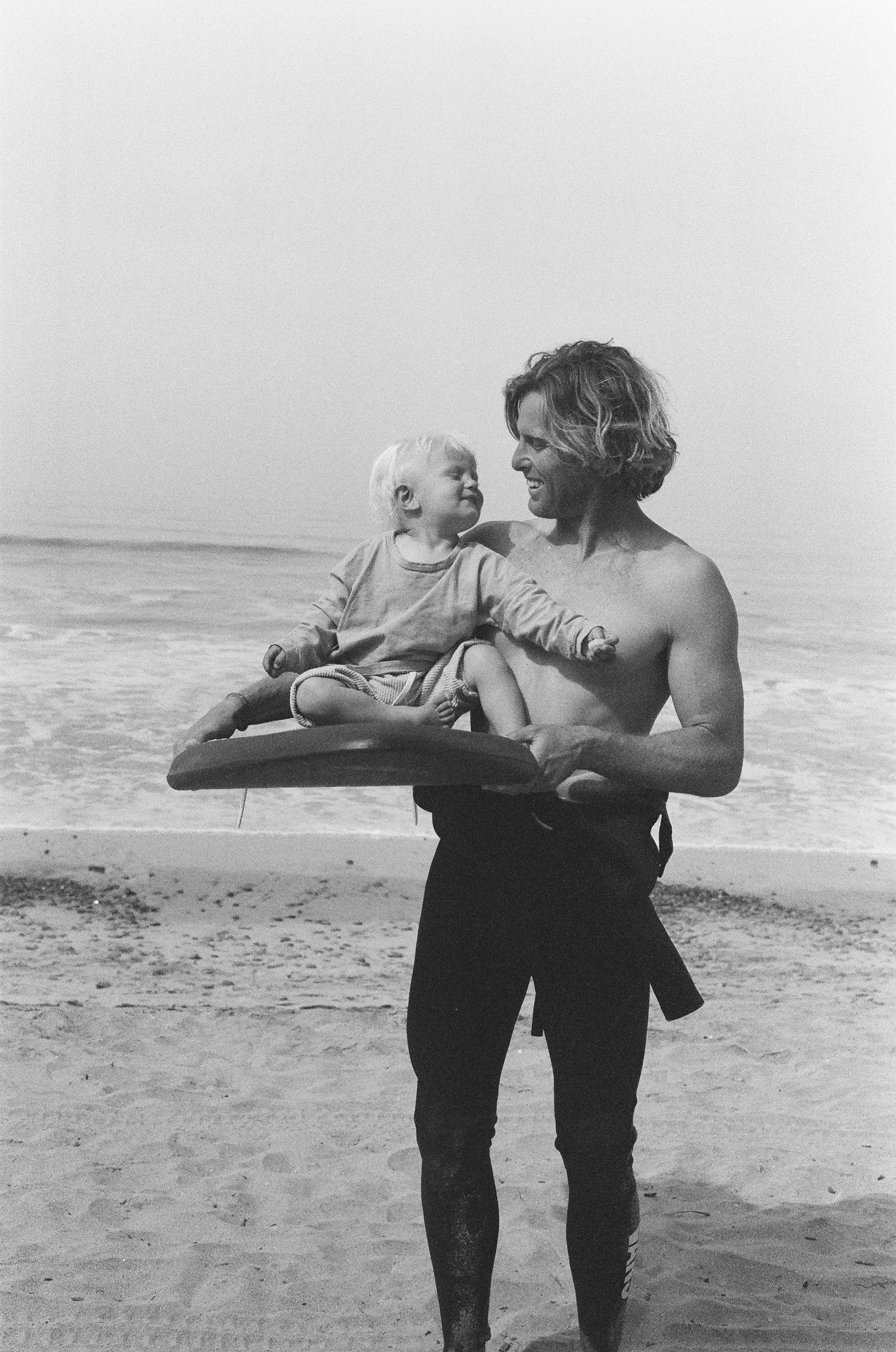 A man on a beach holding a child sitting on a Positive Vibe Warriors surfboard