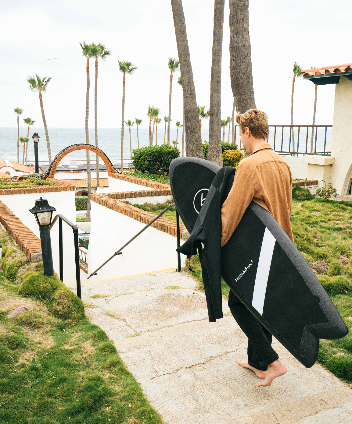 A man walking on a path down to the beach with a black 5 foot 3 inch Formula Fun Foamies Twinnie surfboard.