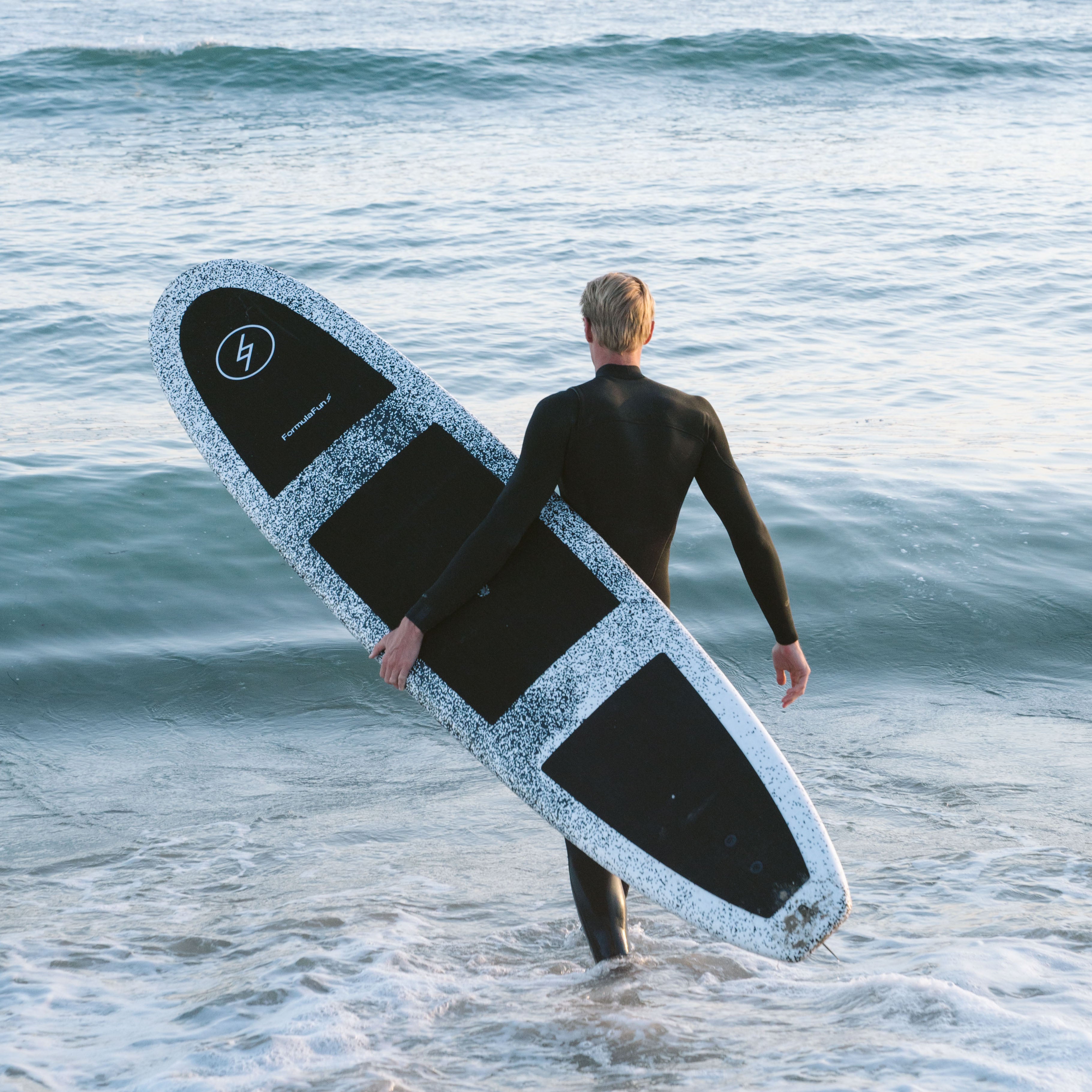 Man in a wetsuit entering the ocean with a speckled Formula Fun Foamies surfboard