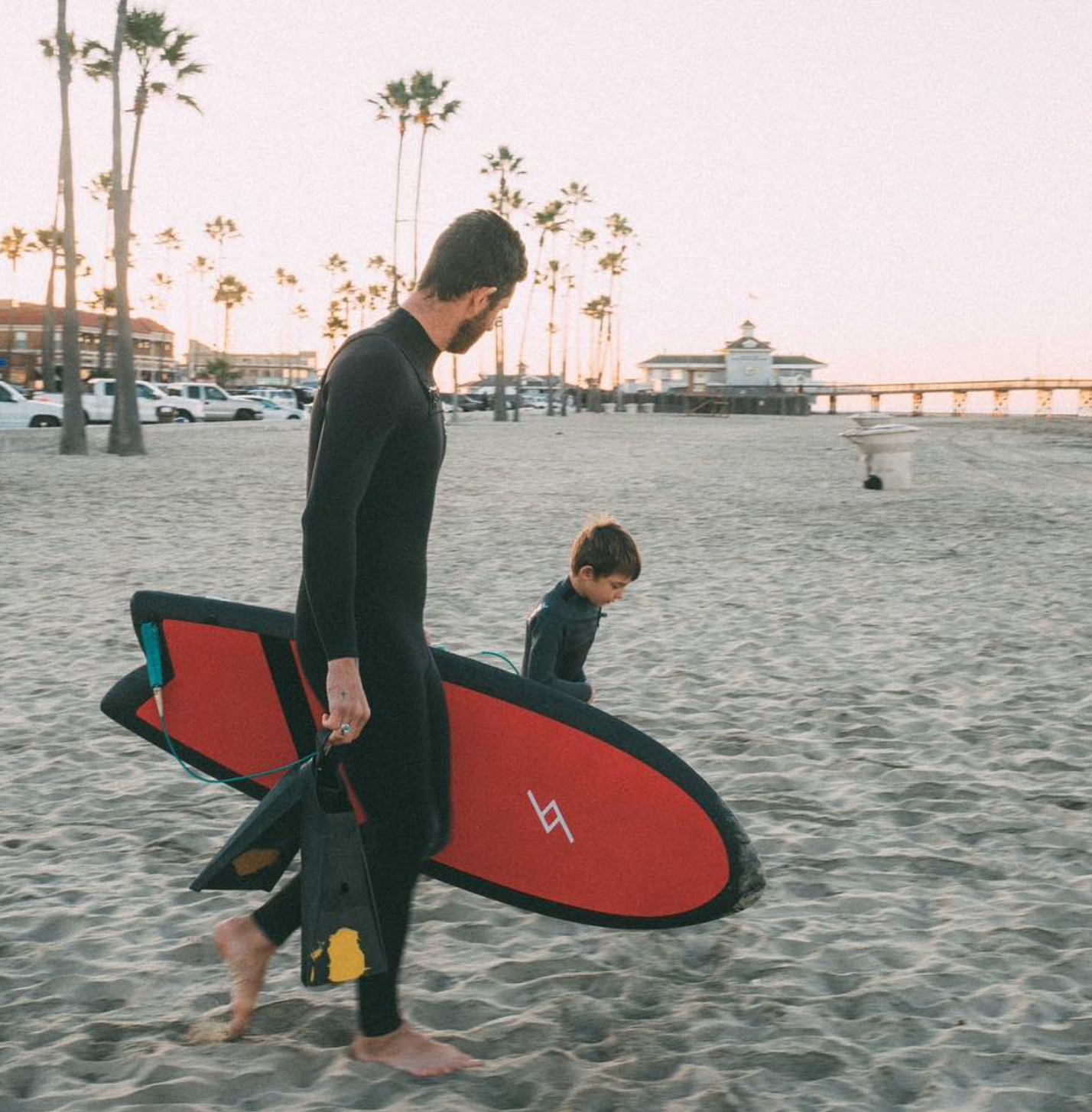 Man in a wetsuit on a beach carrying a red 5 foot 3 inch Formula Fun Foamies Twinnie surfboard walking next to a child