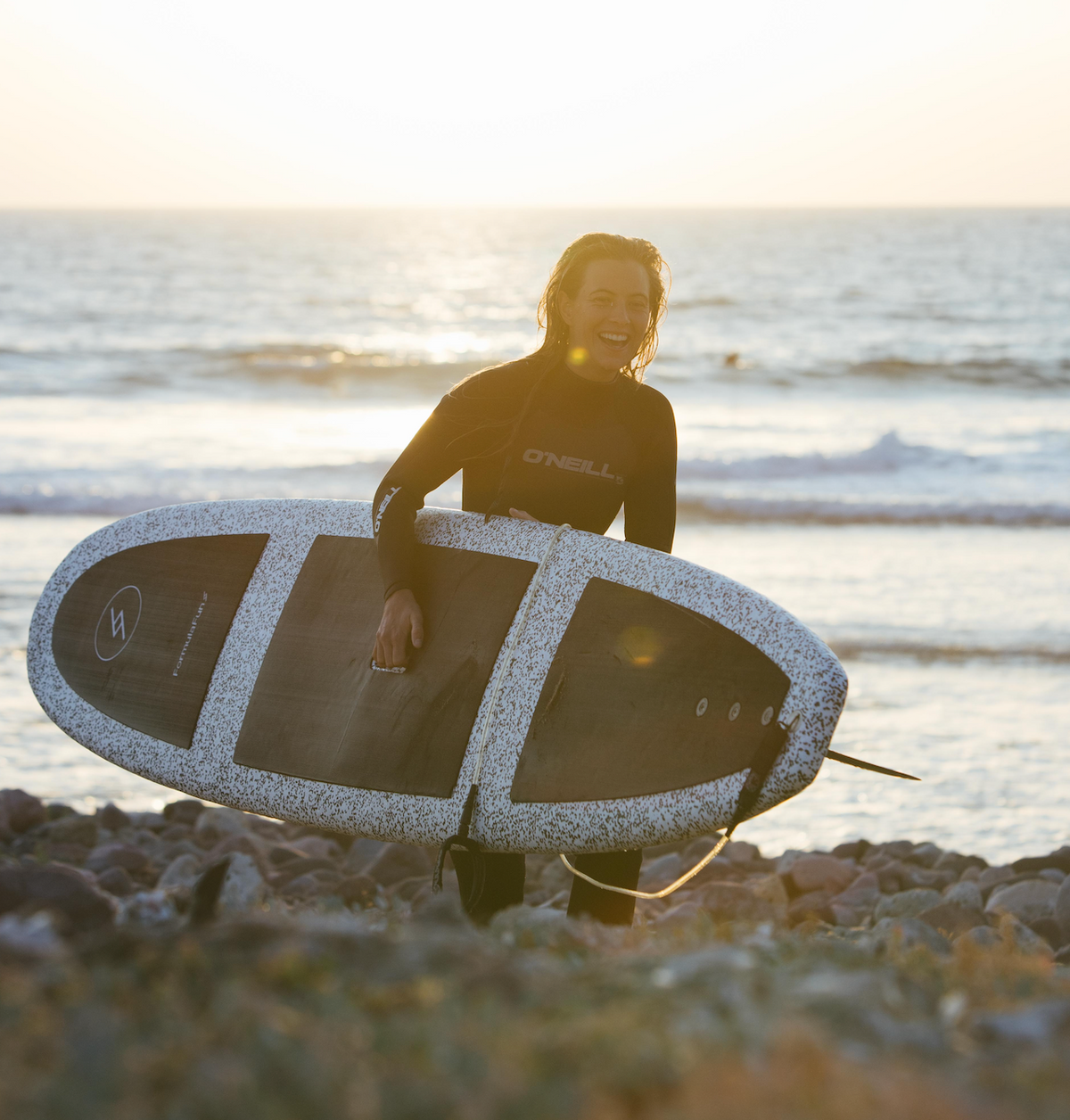 Woman on a beach at sunset carrying a speckled Formula Fun Foamies surfboard