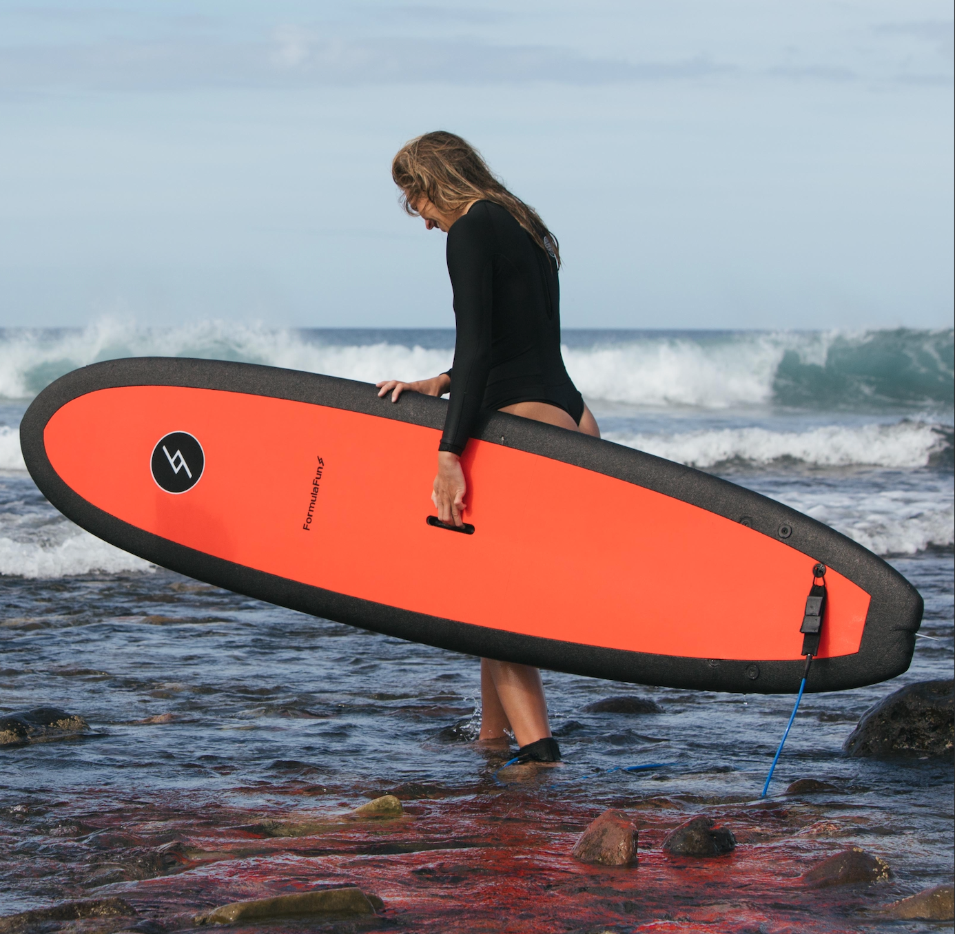 A woman entering the ocean in a wetsuit with a red 7 foot 10 inch Formula Fun Foamies DOHO surfboard