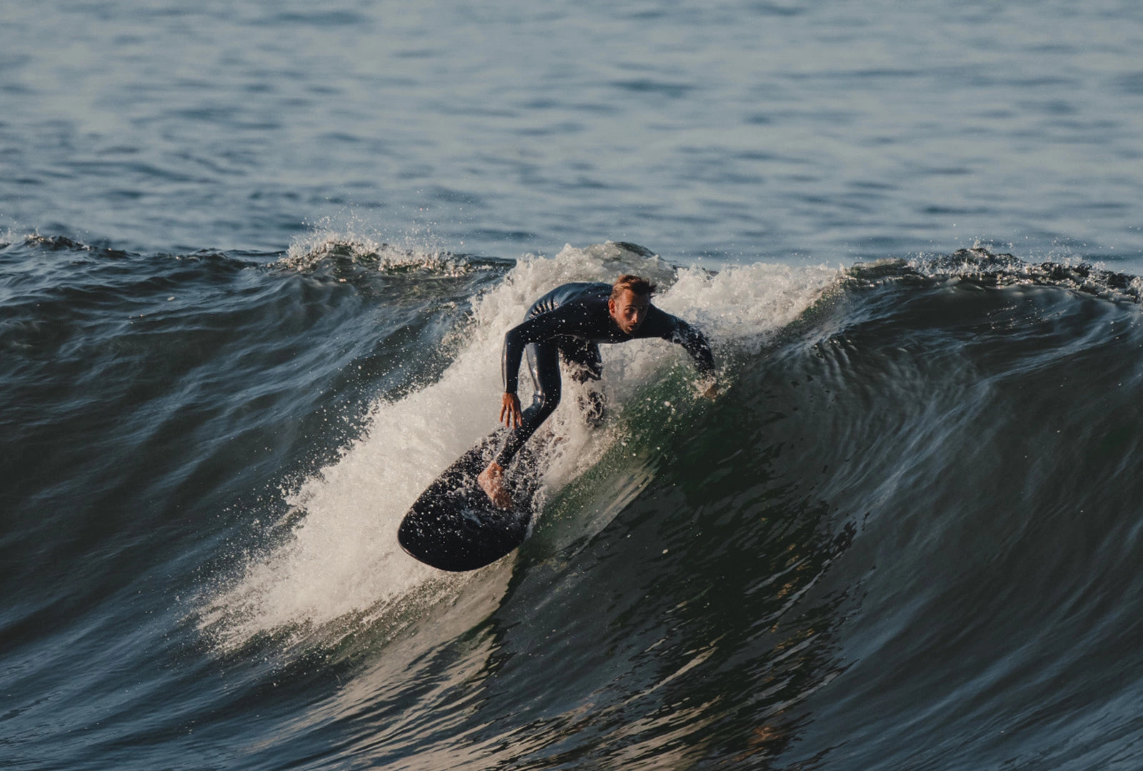 A man in a wetsuit riding a black Almond surfboard in a wave that's breaking next to him