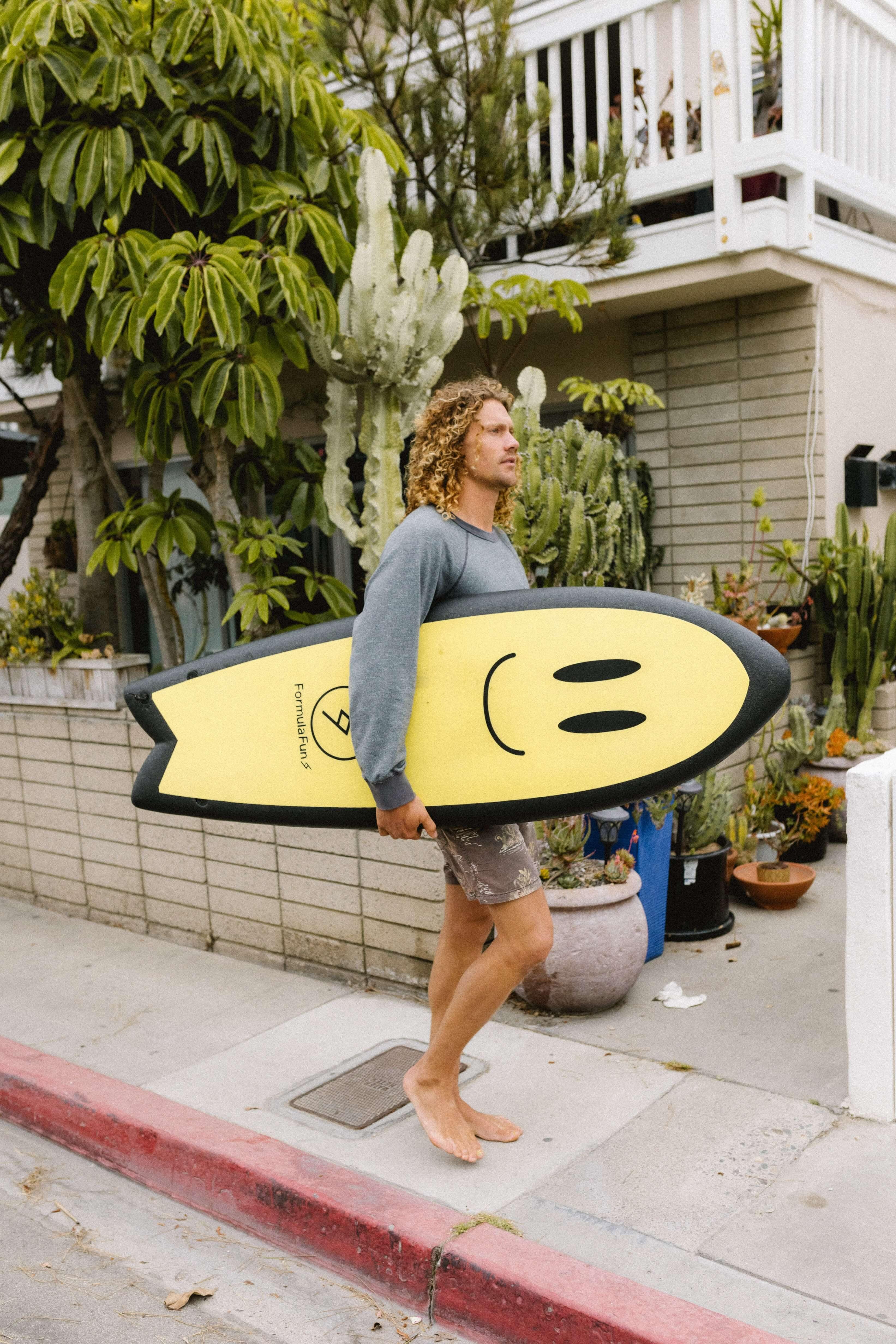 A man walking on a sidewalk with a yellow 5 foot 3 inch Formula Fun Foamies Twinnie surfboard with a smiley illustration