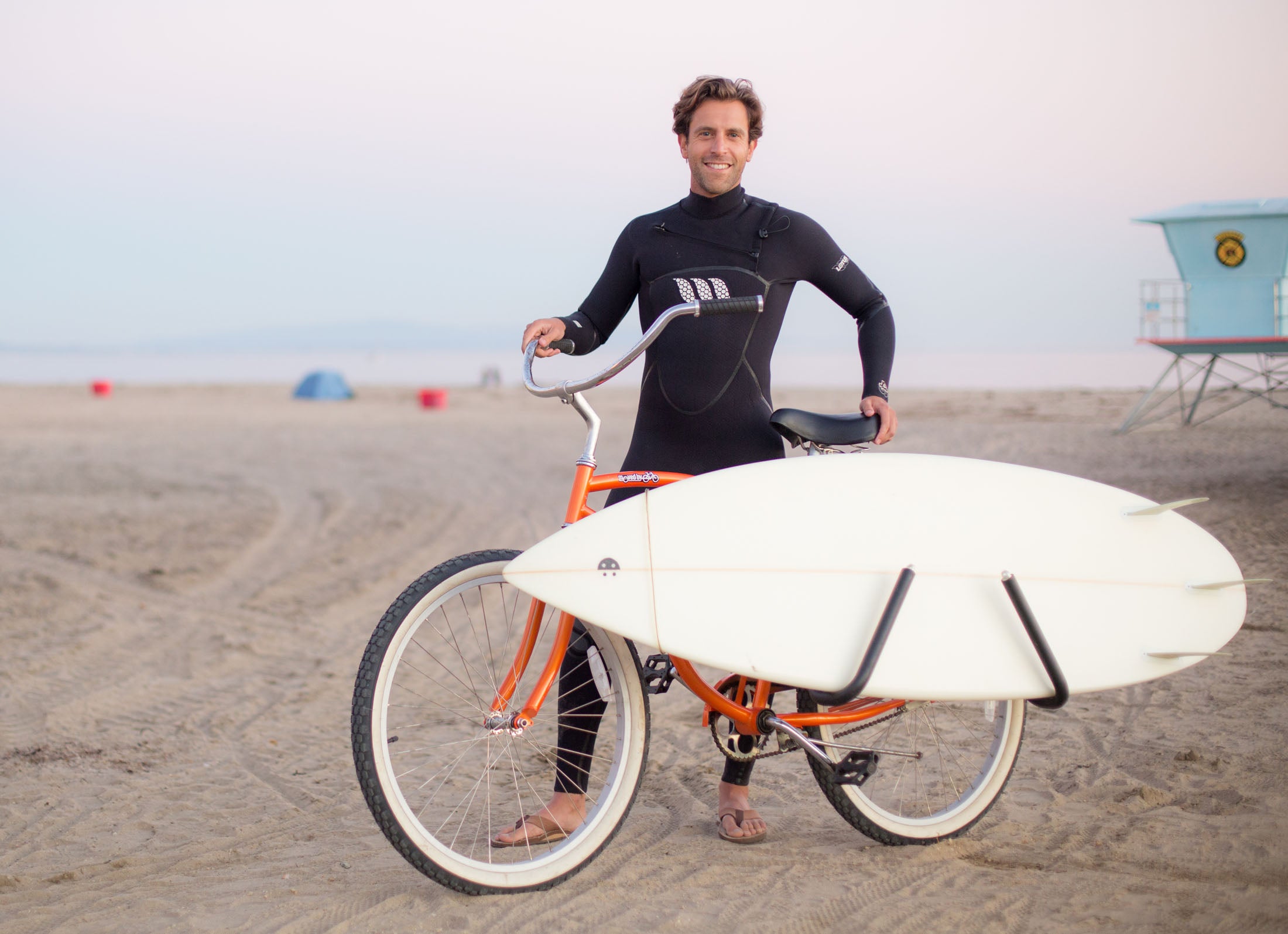 Man on a beach with an orange bike, an MBB shortboard rack carrying a shortboard