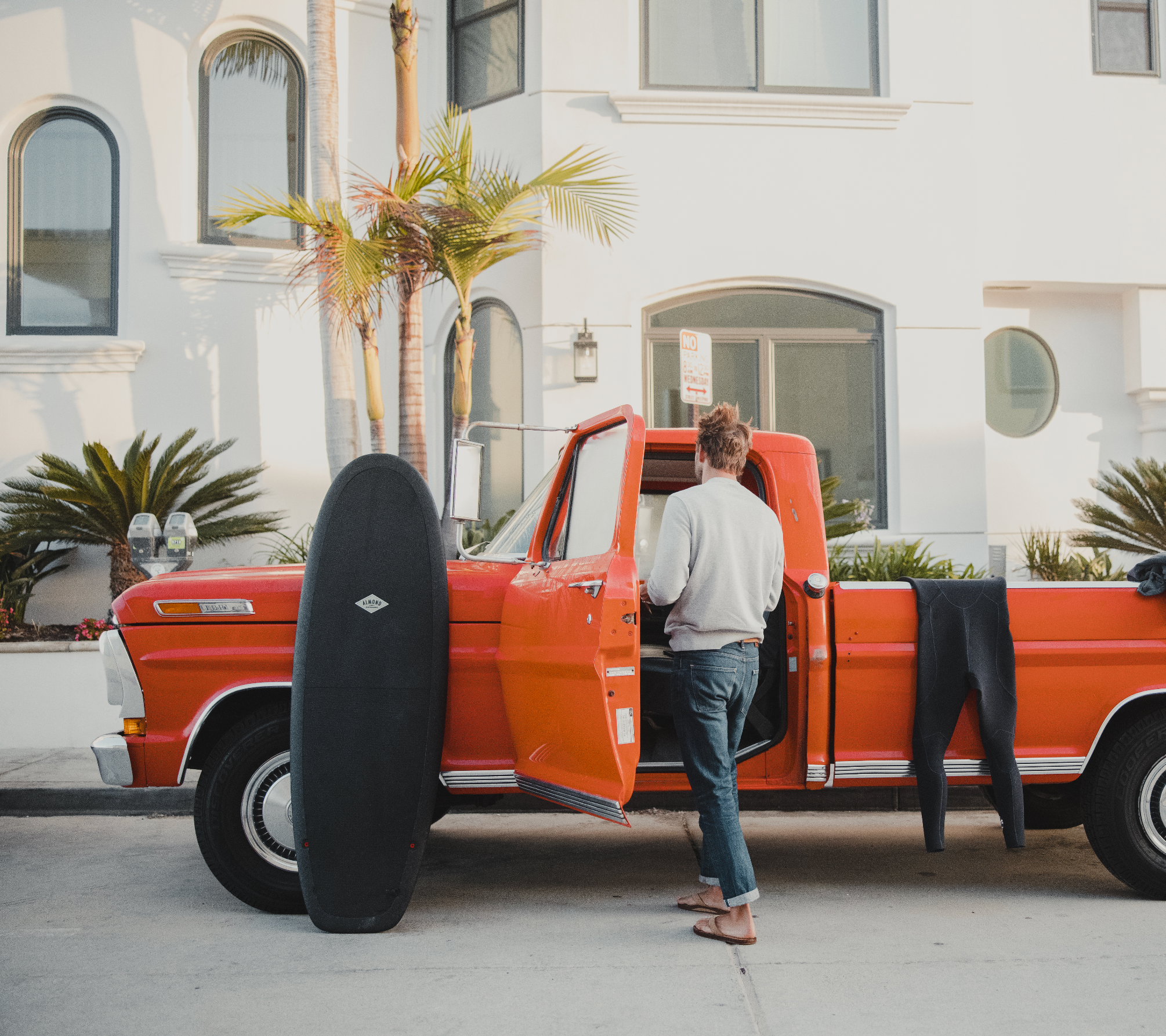 A man entering a red truck with a black 5 foot 4 inch Almond surfboard leaning on the car and a wetsuit